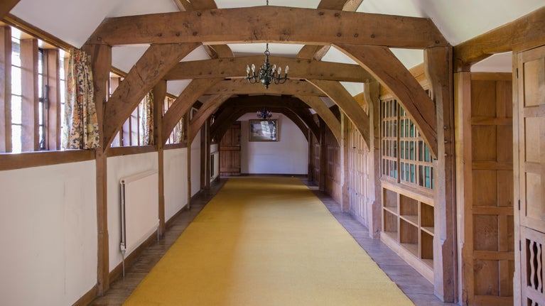 Looking along the first floor gallery at Munstead Wood.  The left wall has a long line of windows, the right is lined with oak cupboards.  The ceiling is crossed by large oak beams and a chandelier hangs in the middle.
